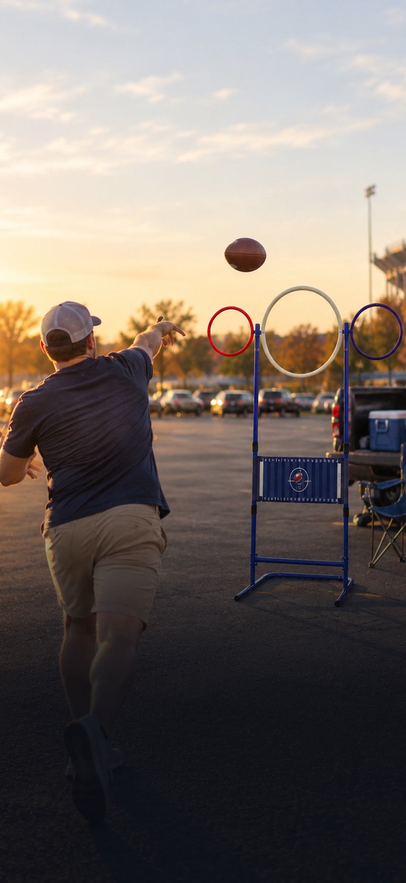 A man mid-throw at a tailgate, a football in the air heading toward the large white centre ring of a QB Rings frame, with a stadium and parked cars behind him at golden hour.